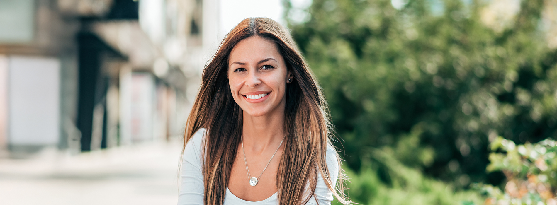 The image shows a woman with long hair smiling at the camera, wearing a white top, standing outdoors during daylight.