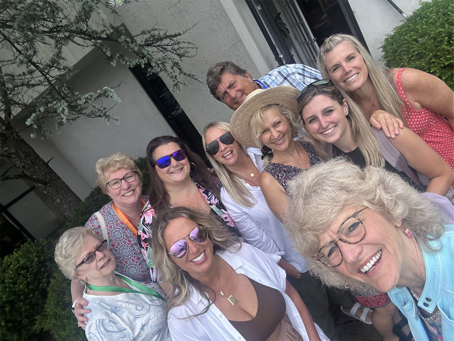The image depicts a group of people posing together outdoors, with some individuals wearing sunglasses and hats, standing in front of a building with trees in the background.