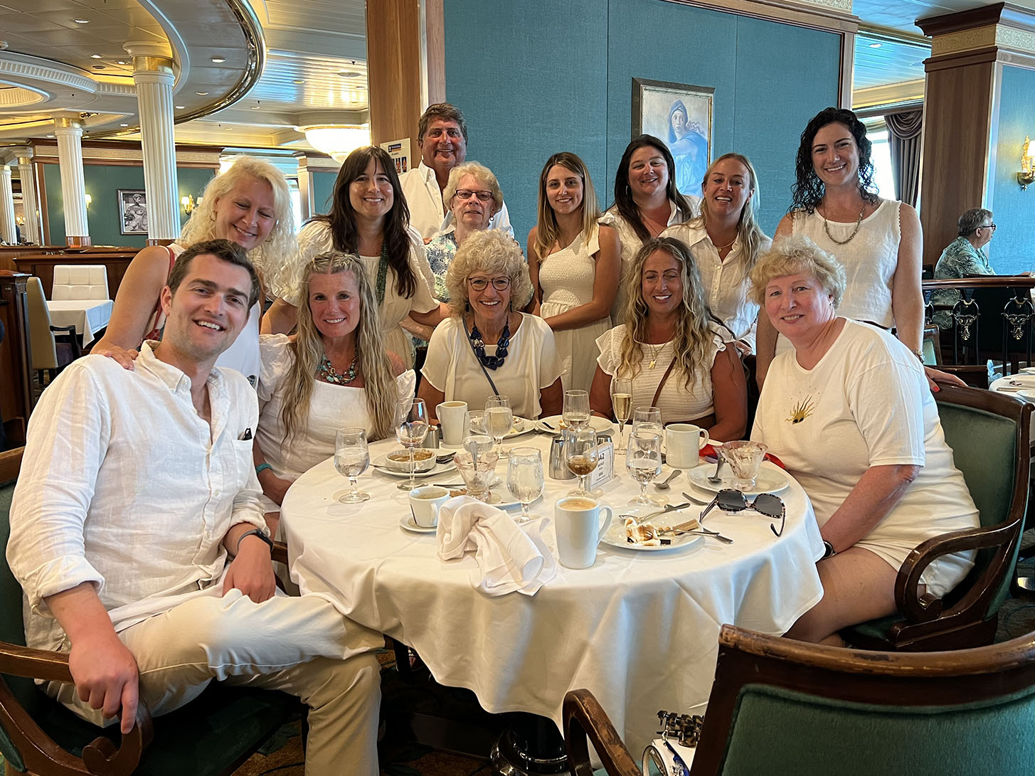 The photo shows a group of people wearing white attire gathered around a dining table at an event, posing for the camera with smiles.