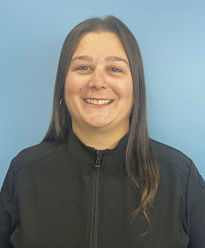 The image shows a woman standing against a blue background with a white stripe at the bottom. She has long brown hair, is wearing a black jacket over a lighter-colored shirt, and appears to be smiling.