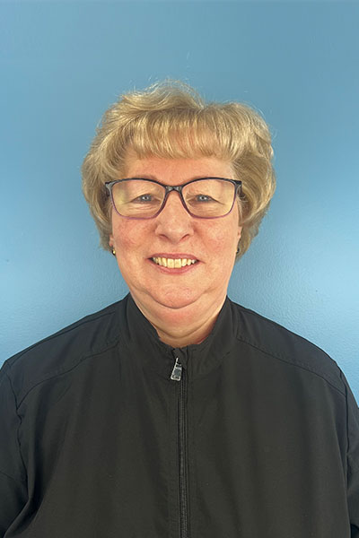 The image shows a person wearing glasses, with short hair, posing for a portrait against a blue wall. They are wearing a dark jacket with a white collar and a name tag that reads  Coach.