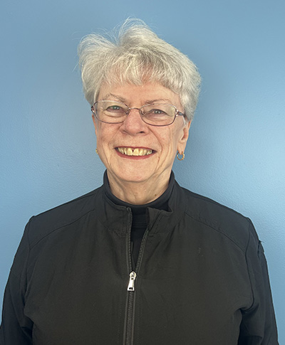 An elderly woman with white hair wearing glasses, a black jacket, and a smile poses for a portrait against a blue background.