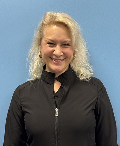The image shows a woman posing for a portrait with a neutral background. She is smiling at the camera, wearing a black jacket over a long-sleeved shirt, and has short blonde hair.