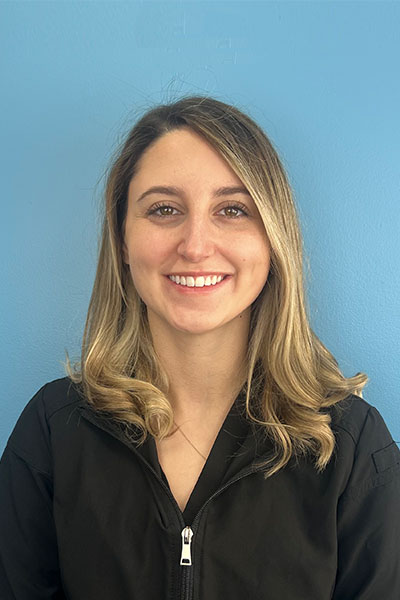 The image shows a woman with short hair smiling at the camera, wearing a black jacket with a white zipper, positioned against a blue background.