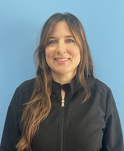The image is a portrait of a woman standing against a blue background. She has long hair, is wearing a black jacket with a zipper, and has her mouth slightly open.