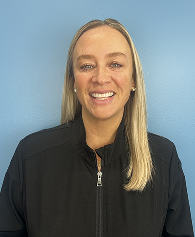 The image shows a woman smiling at the camera  she has light-colored hair, wears a dark jacket, and is standing against a blue background.