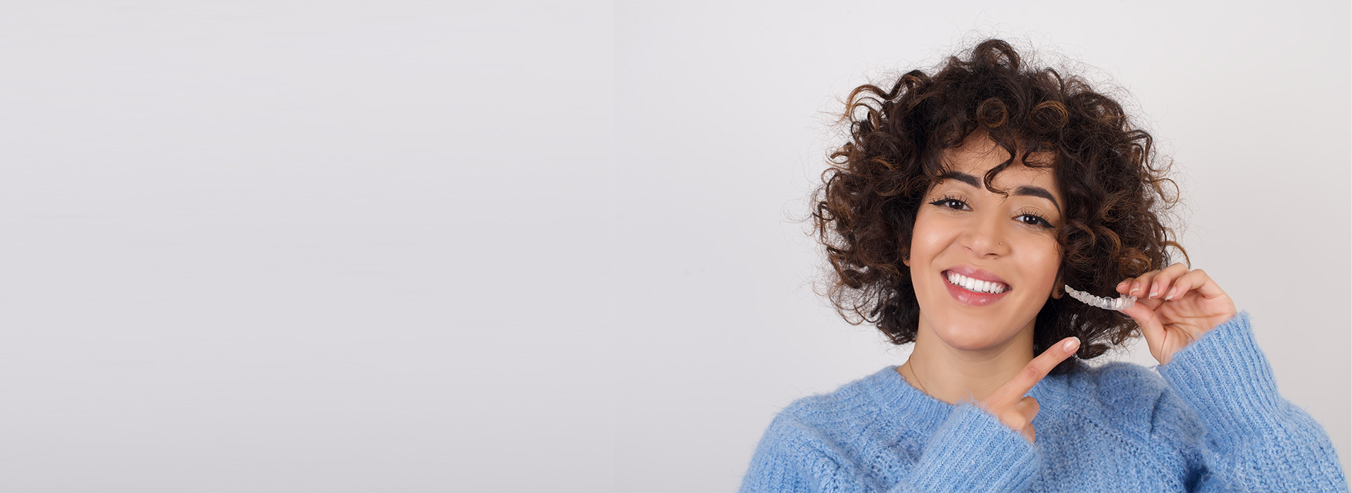 The image features a person holding a toothbrush with curly hair, smiling at the camera, against a white background.