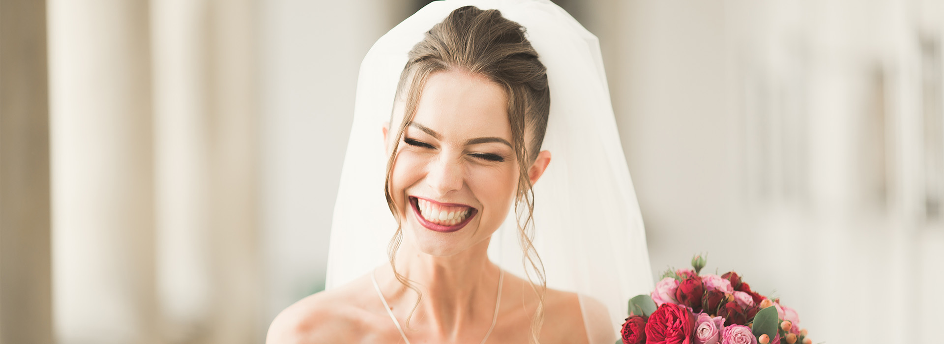 Woman wearing a white wedding dress with veil, smiling and looking upwards, standing next to a bouquet of flowers, in front of an archway.