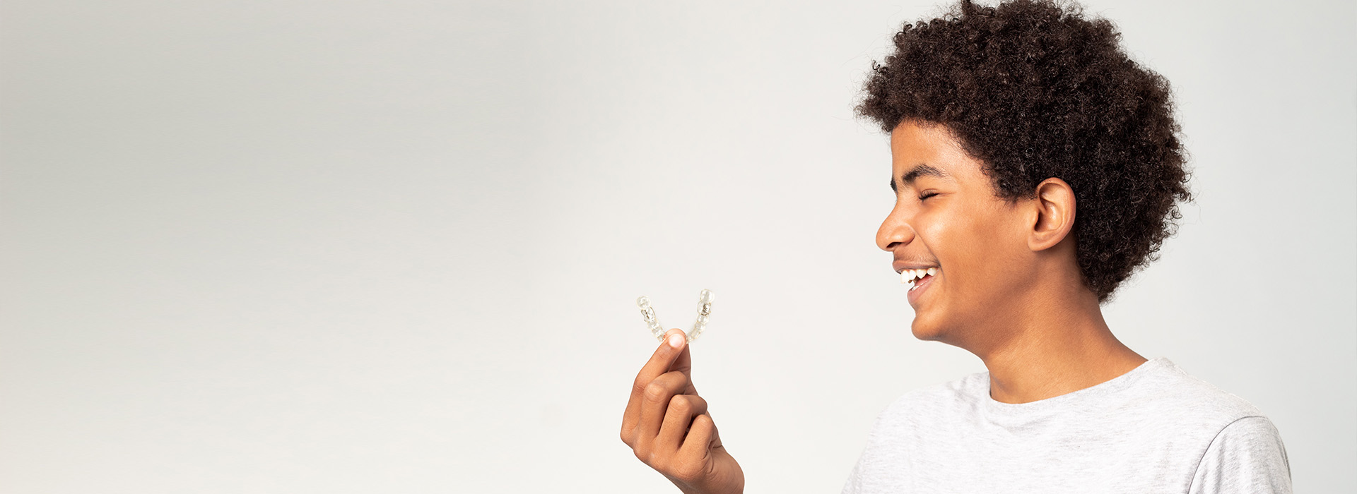 This is a photograph featuring a person with curly hair smiling at the camera while holding an object, possibly a key or a small toy, against a plain background. The individual appears to be a young adult wearing a light-colored top and has a relaxed expression.
