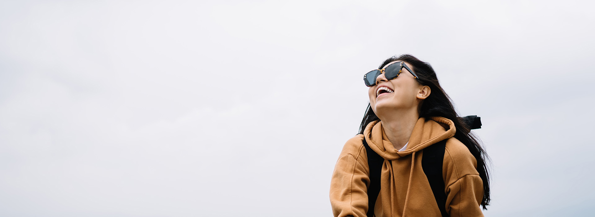 The image shows a person with sunglasses on their head, facing away from the camera, standing outdoors with a clear sky background, wearing a backpack and a hoodie, looking towards the horizon or something beyond the frame of the photo.