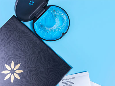 The image shows a dark-colored box with a light blue lid containing a small amount of green substance, placed on a surface along with an open booklet displaying a patterned design and a blue plastic cup with a logo.