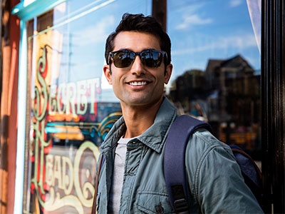 The man in sunglasses is standing in front of a storefront with his hands in his pockets, wearing a backpack.