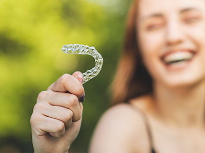 The image shows a person holding up a toothbrush with a smiley face on it against a blurred outdoor background, indicating good oral hygiene practices.