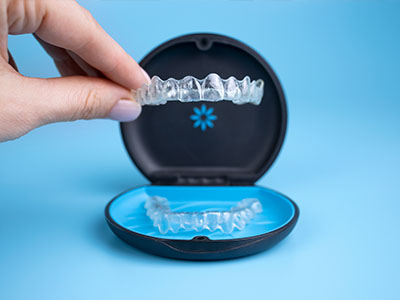 The image shows a person s hand holding a clear plastic tray with a set of transparent braces inside, placed on a blue background next to an open case containing more of the same type of braces.
