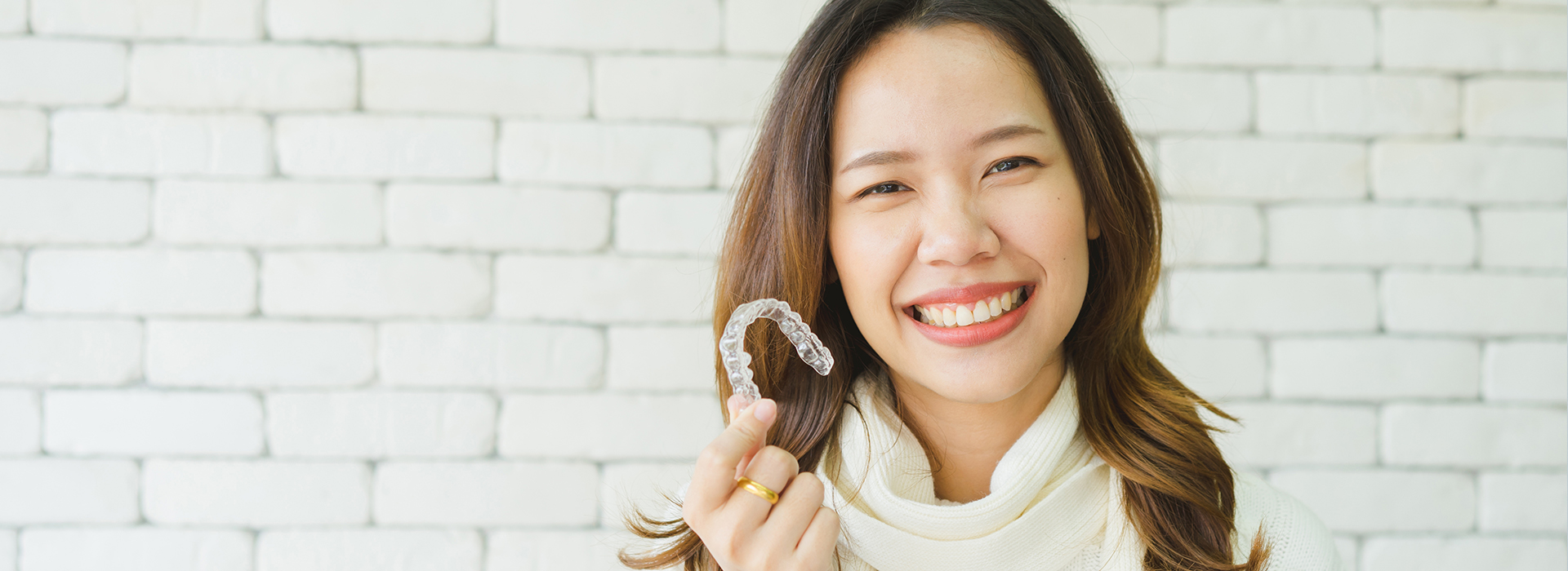 A woman with a smile holds up a toothbrush in front of her face, standing against a brick wall background.