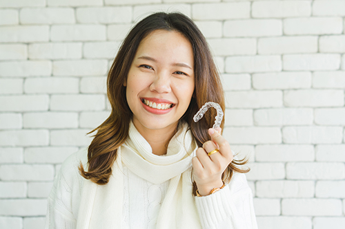 A smiling woman holding a white object with her left hand, standing against a brick wall background.